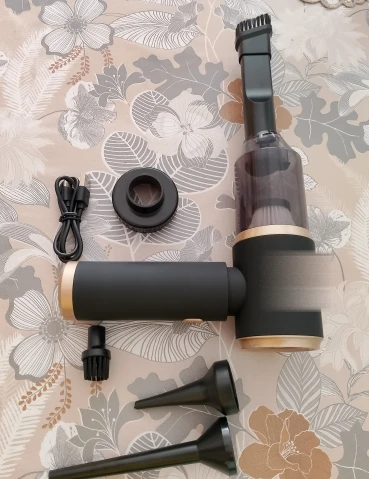 Komplet rozłożony na kwiecistej tkaninie: odkurzacz z założoną szczotką i przezroczystym pojemnikiem, obok kabel USB-C, adapter-kubek, szczotka okrągła i dwie stożkowe końcówki.